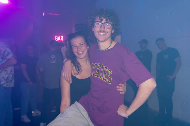 Two partygoers posing in a purple-lit, hazy club with a neon bar sign behind them.