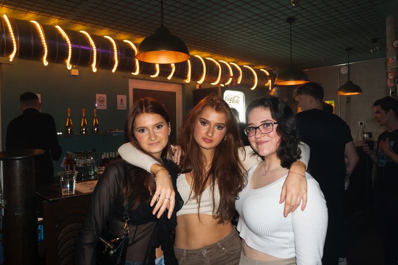 Three friends smiling in a bar with warm orange tube lights above.